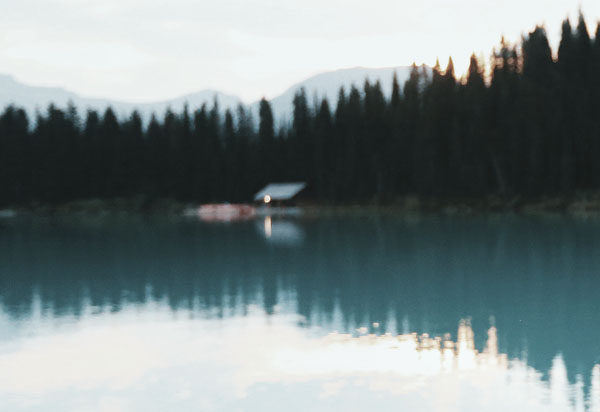 Scandinavian scenic shot, soft focus, foreground lake with reflection of tall pine trees and mountain in background, cabin on edge of like with external light